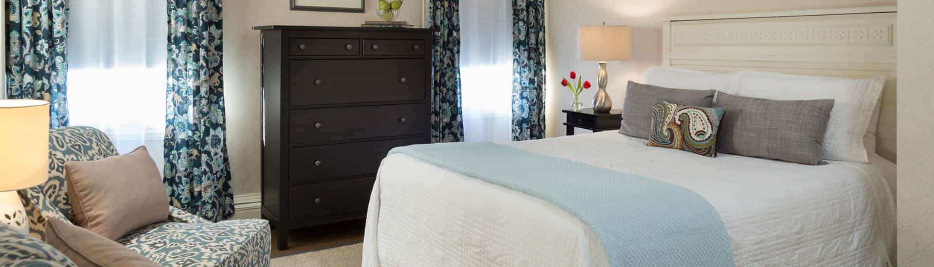 Bedroom with hardwood flooring, white bedding, dark wooden dresser, and upholstered armchairs