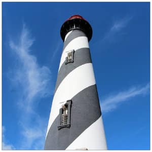 View from the bottom of a large white and black striped lighthouse against a bright blue sky