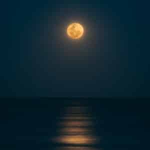 Bright yellow, orange moon in the middle of a night sky lighting up over calm water