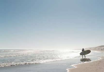 Pristine beach with a lone surfer standing at the shore and blue skies overhead