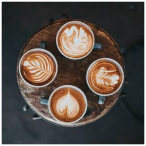 Four black mugs of lattes with special foam designs sitting on a brown wooden stool.