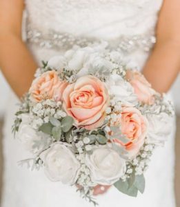A bride in a white dress holding a bouquet or white and peach roses and baby's breath