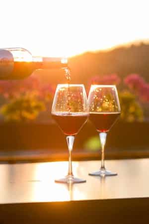 A bottle pouring red wine into two glasses with vineyard and glowing sunset in background 