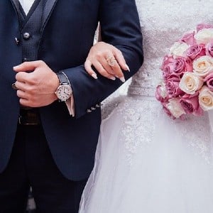 Bride in white dress holding wedding bouquet with arm linked into man in black tuxedo