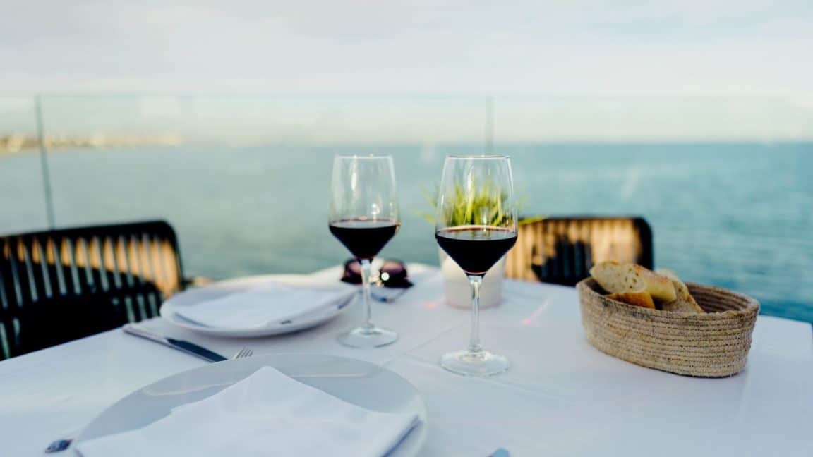 Table set for an elegant meal with two glasses of red wine, basket of bread, and view of ocean