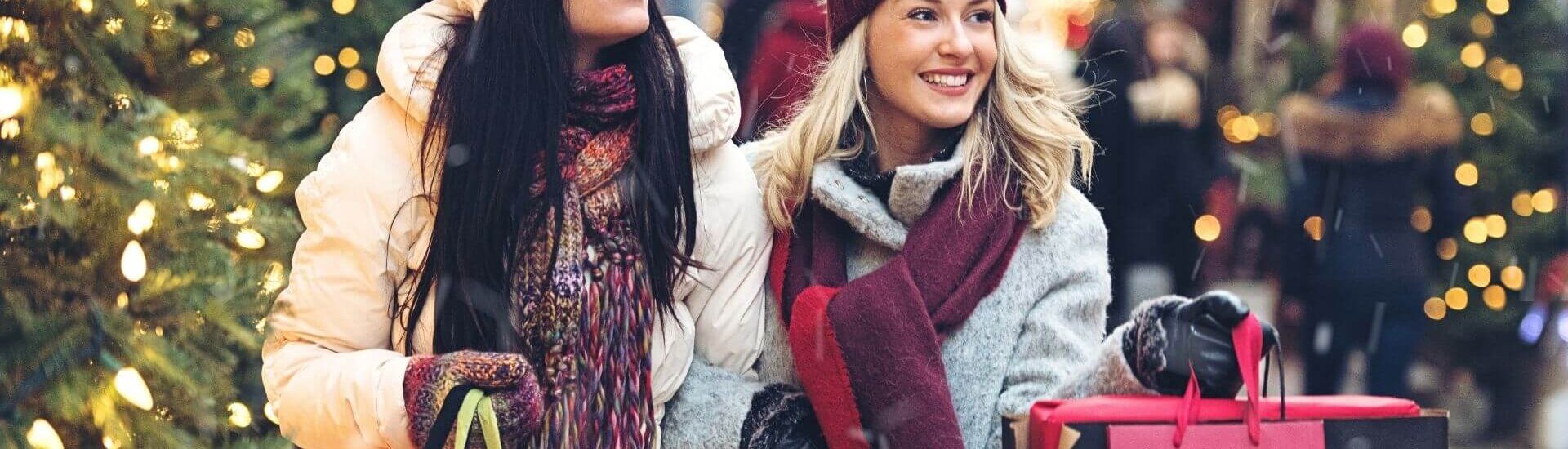 two women in winter coats and hats smile while carrying shopping bags on a decorated street lined with christmas lights and trees