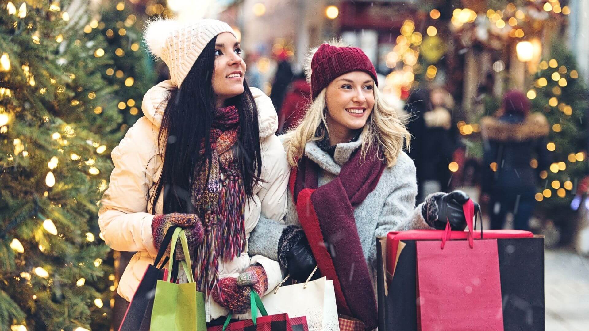 two women in winter coats and hats smile while carrying shopping bags on a decorated street lined with christmas lights and trees
