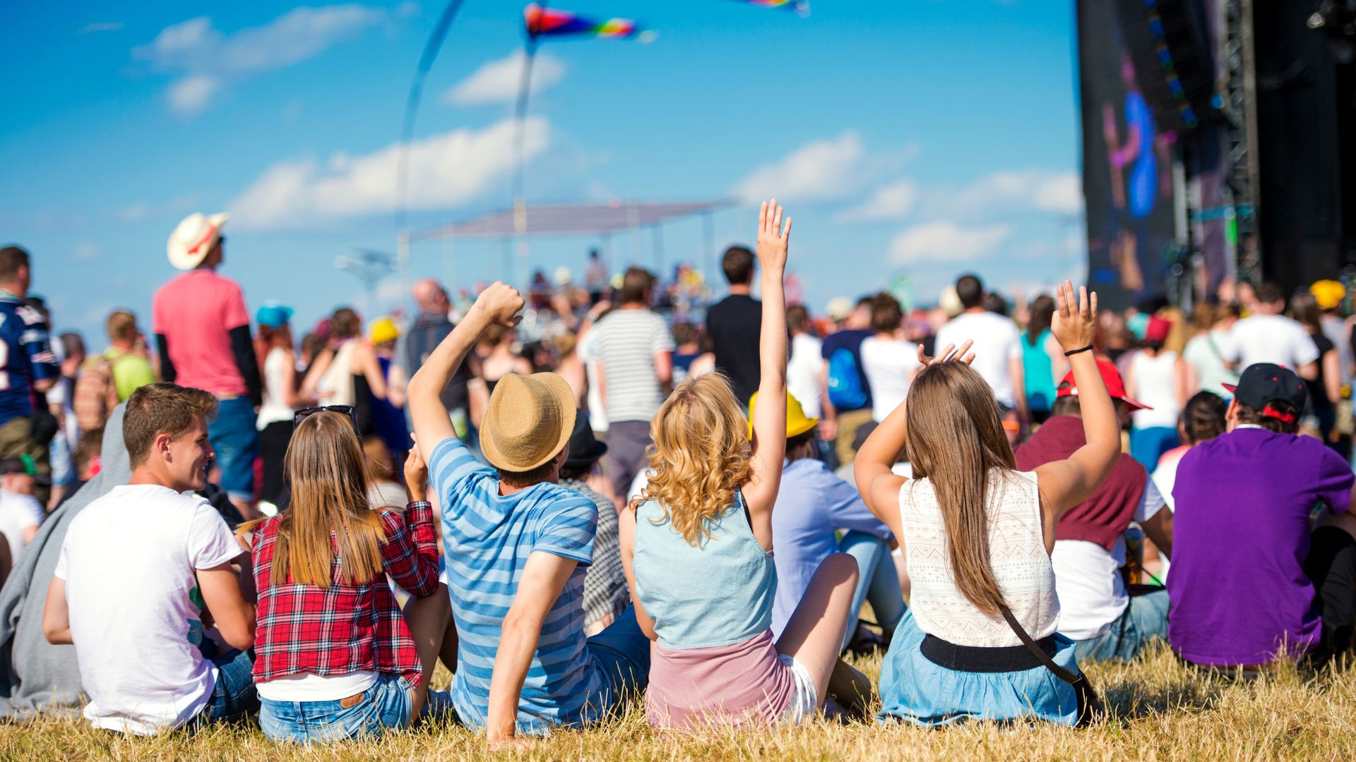 A group of people sitting in the grass at a music festival in a park.