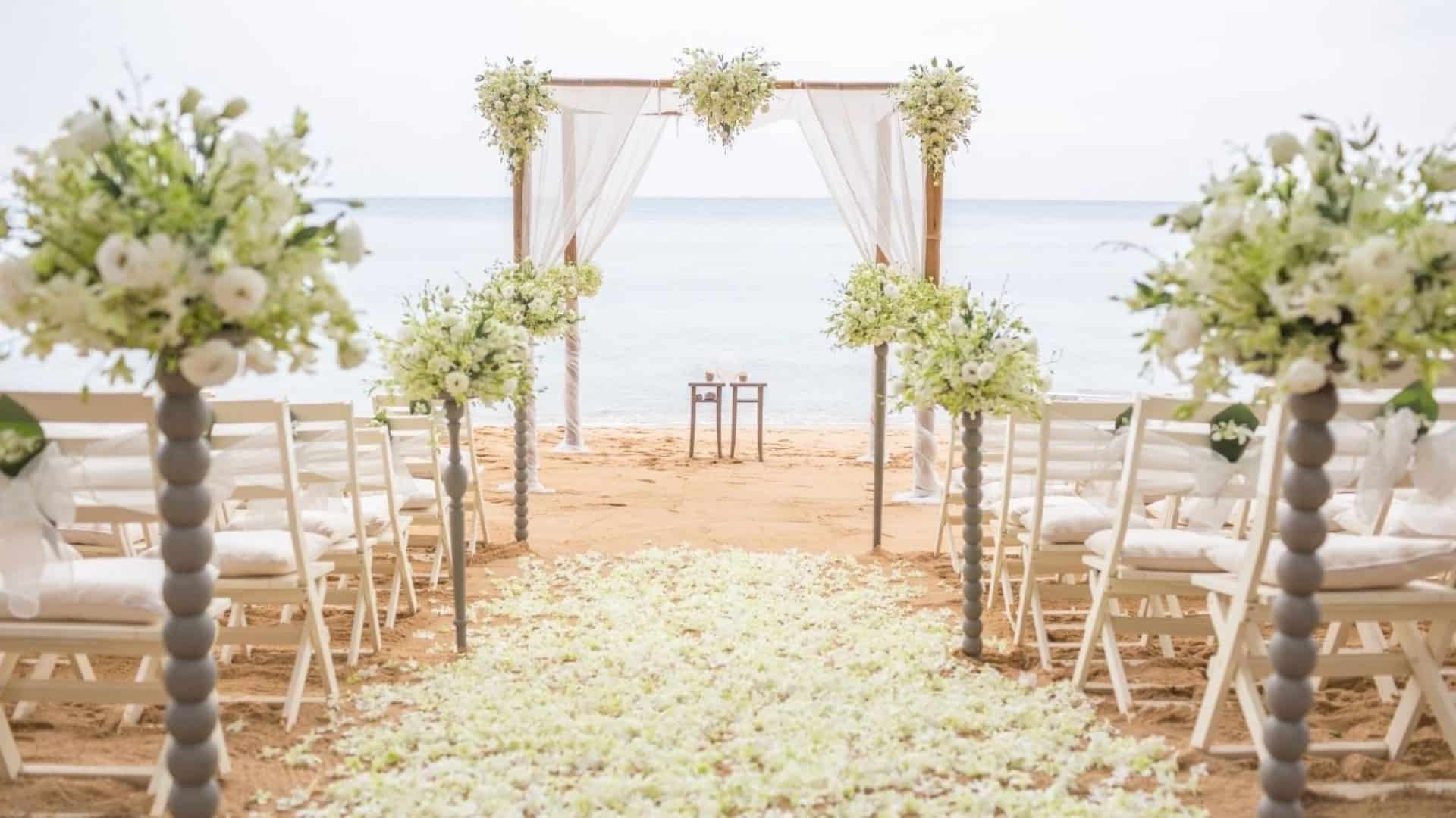 Beach wedding set up with chairs, flowers, petals in the aisle, and a floral decorated canopy at the water’s edge