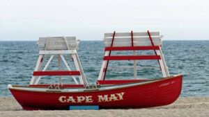 life boat resting on the beach behind lifeguard seats. text painted on the side of the boat: “cape may.”