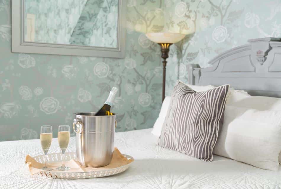 White tray with two glasses filled with Champagne and a bottle of wine chilling in a silver bucket sitting on top of a bed with white bedding
