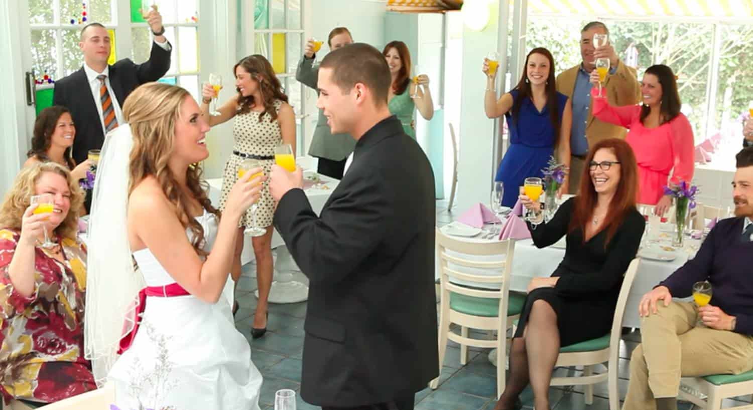 Bride and groom smiling and toasting each other with people in the background raising their glasses