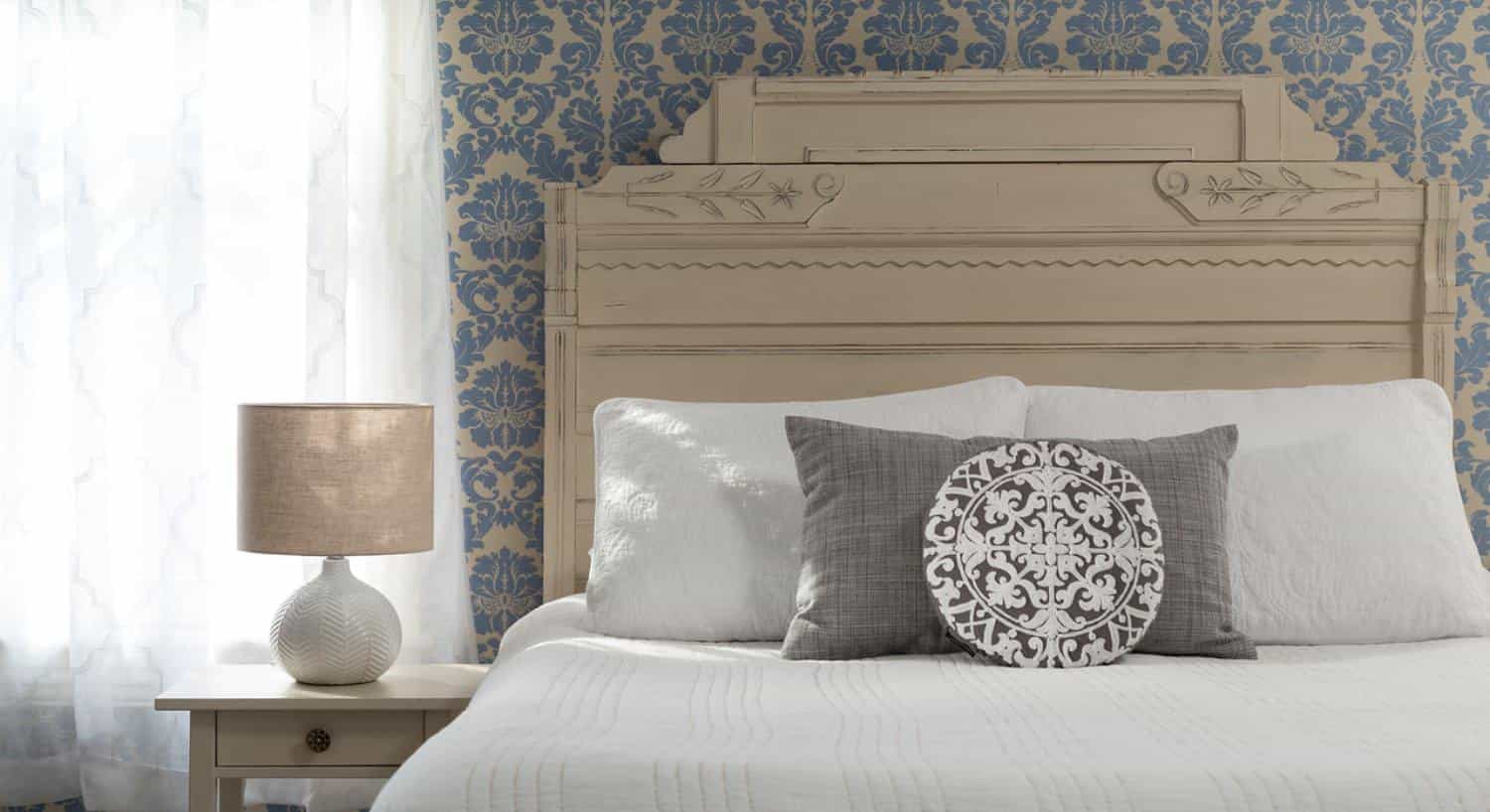 Bed with antiqued headboard, white bedding, gray pillow, brocade wallpaper, and window with white opaque curtains