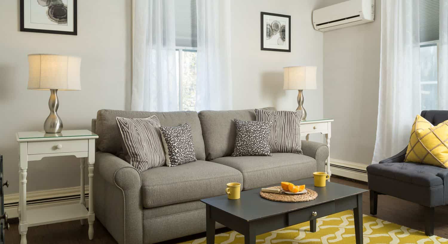 Sitting area with gray upholstered coach, gray and white and yellow pillows, hardwood flooring, light walls, dark wooden coffee table and yellow and white area rug