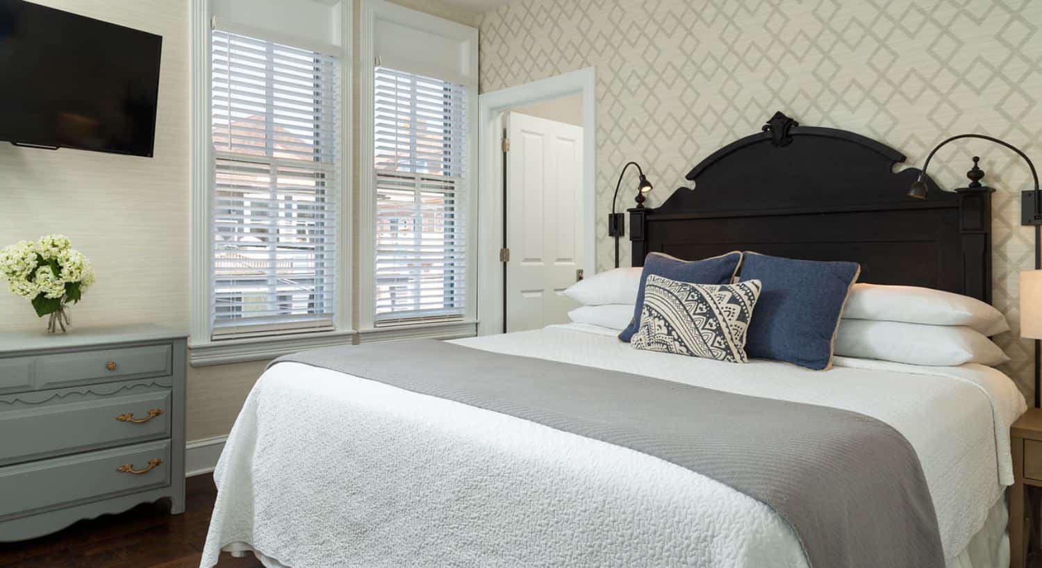 Bedroom with dark wooden headboard, hardwood flooring, light colored wallpaper, white bedding, gray dresser, and flat-screen TV mounted on the wall