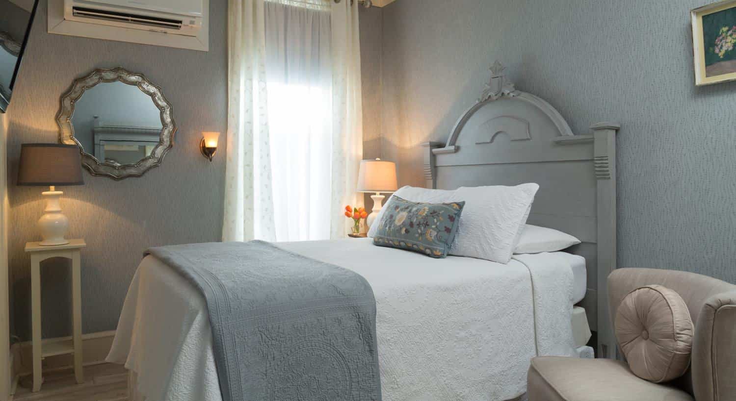 Bedroom with gray wooden headboard, white bedding, hardwood flooring, light gray patterned wallpaper, small upholstered chair, and mirror on the wall