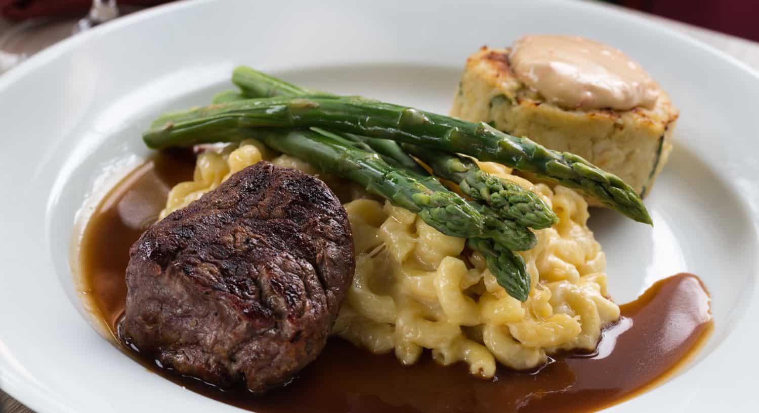 Close up view of a juicy steak, creamy macaroni and cheese, asparagus, and crab cake on white porcelain plate