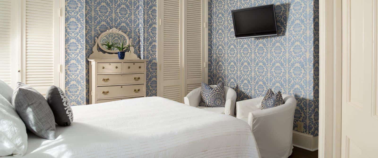 Bedroom with white bedding, hardwood flooring, brocade blue wallpaper, sitting area with white slipcovered chairs, and wall-mounted flat-screen TV