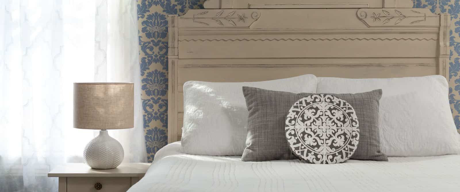 Bedroom with light-colored ornate headboard, white bedding, brocade blue wallpaper, and light-colored nightstand