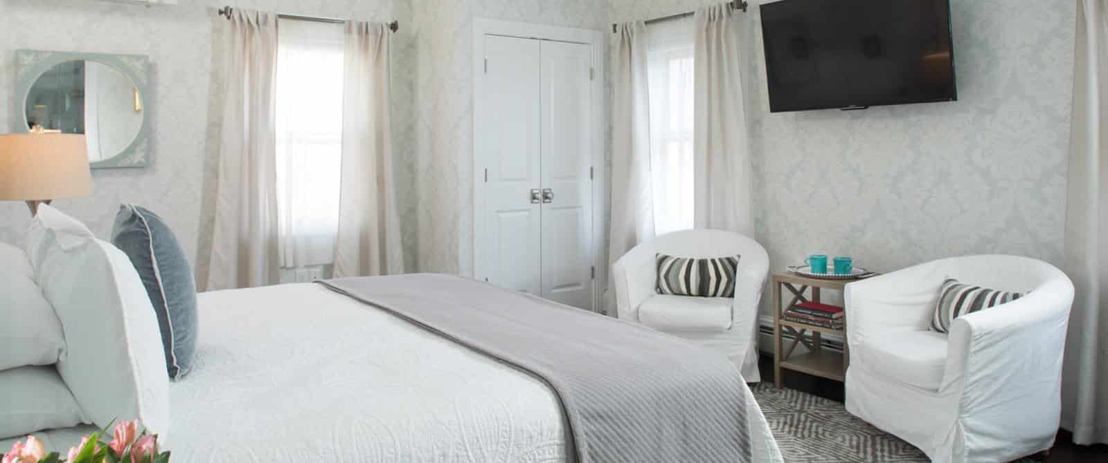 Bedroom with white bedding, gray blanket, hardwood flooring, light brocade wallpaper, sitting area with white slip-covered chairs, and wall-mounted flatscreen TV