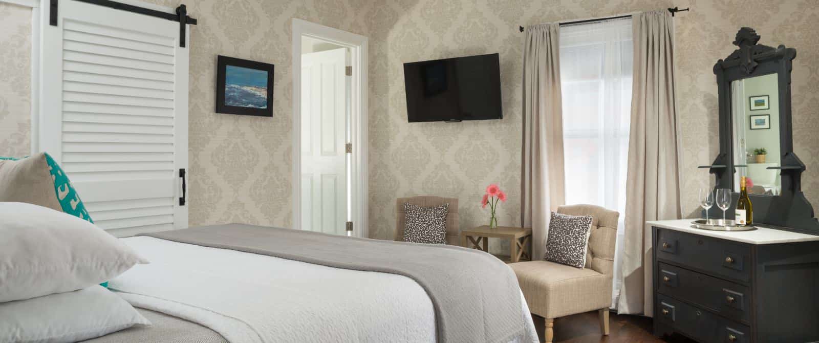 Large bedroom with white bedding, gray blanket, sitting area with tan upholstered chairs, dark wooden dresser with mirror, and wall-mounted TV