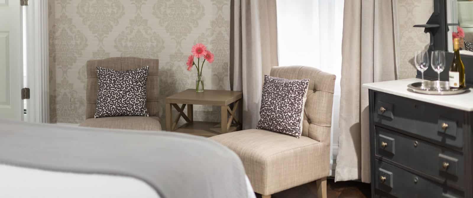 Large bedroom with white bedding, gray blanket, sitting area with tan upholstered chairs, and dark wooden dresser with mirror