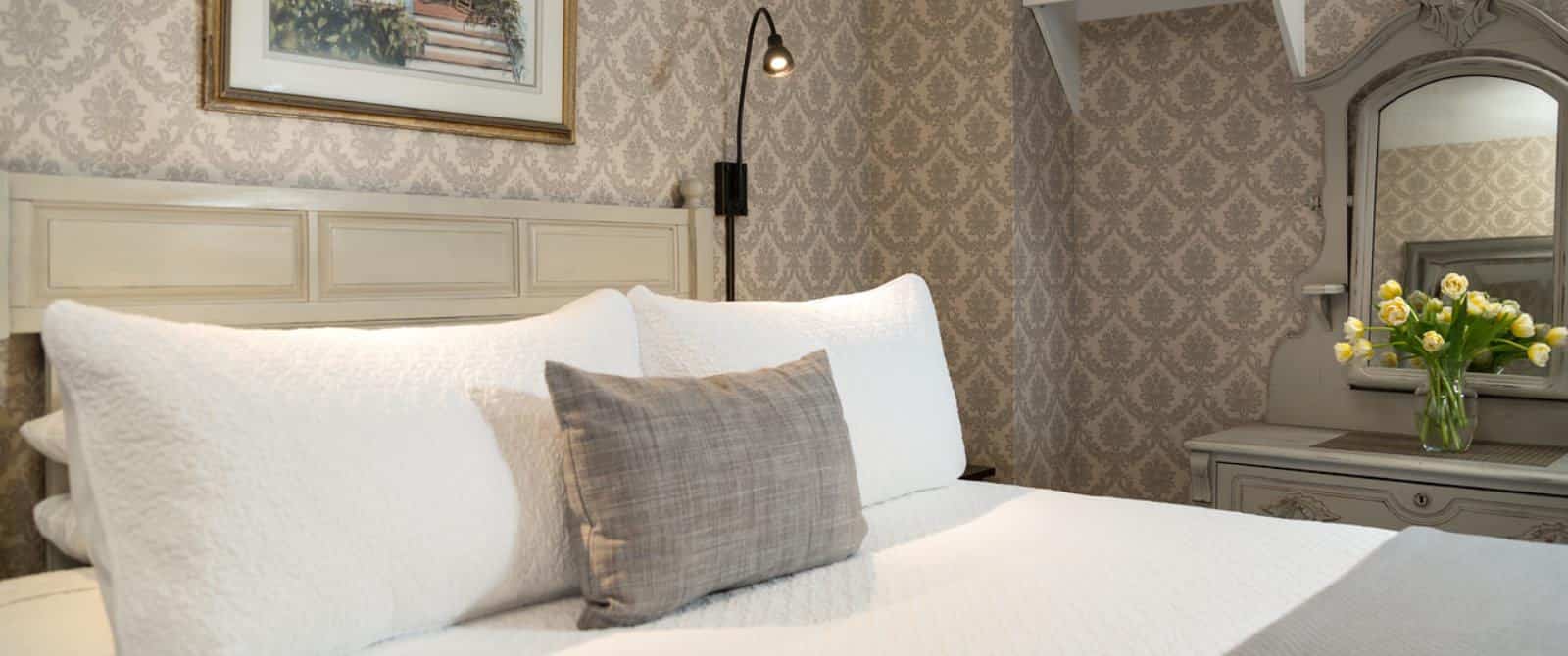 Bedroom with light-colored wooden headboard, white bedding, gray blanket, and gray ornate dresser with mirror