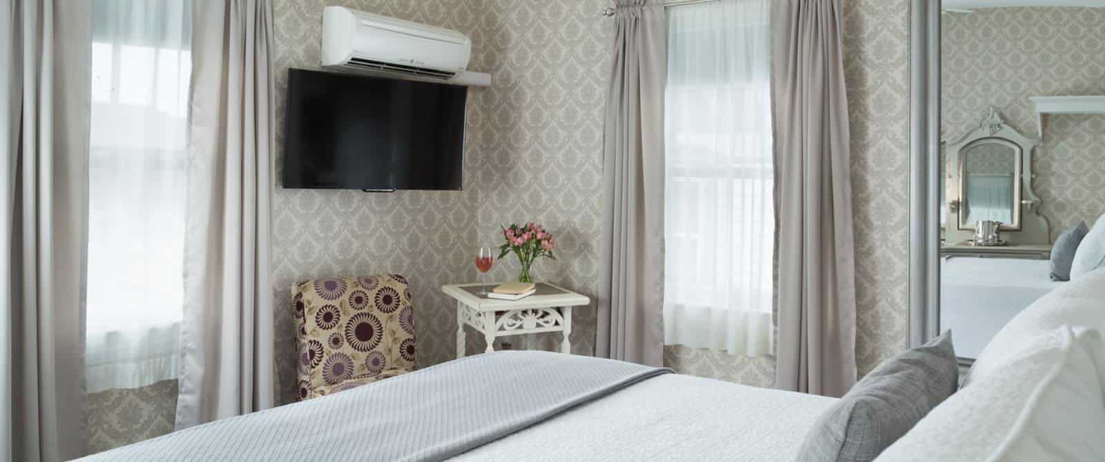 Bedroom with light-colored wooden headboard, white bedding, gray blanket, sitting area with upholstered chair, large mirror, and wall-mounted flat-screen TV