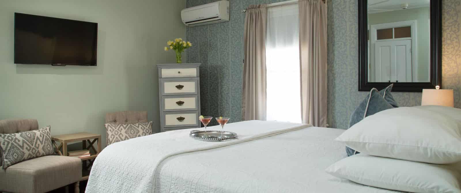 Bedroom with white bedding, hardwood flooring, sitting area with upholstered chairs, and wall-mounted flat-screen TV