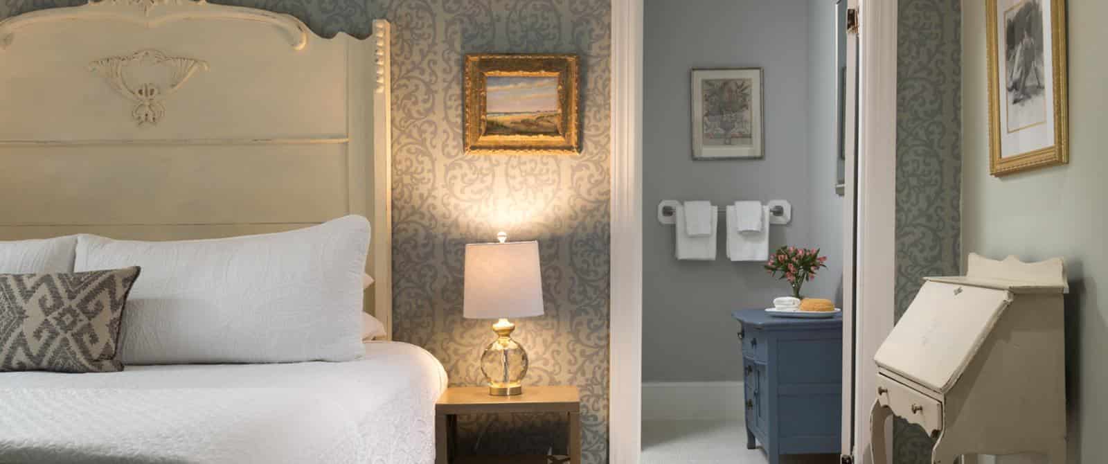 Bedroom with large light-colored ornate headboard, white bedding, and view into attached bathroom with blueish-gray cabinet
