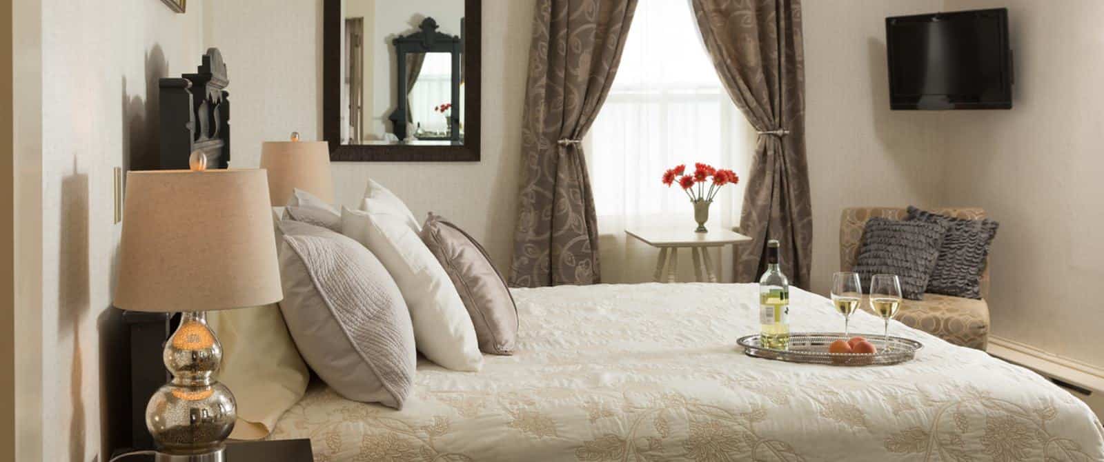 Bedroom with dark wooden ornate headboard, cream-colored bedding, dark wooden nightstand, upholstered chair, and wall-mounted flat-screen TV