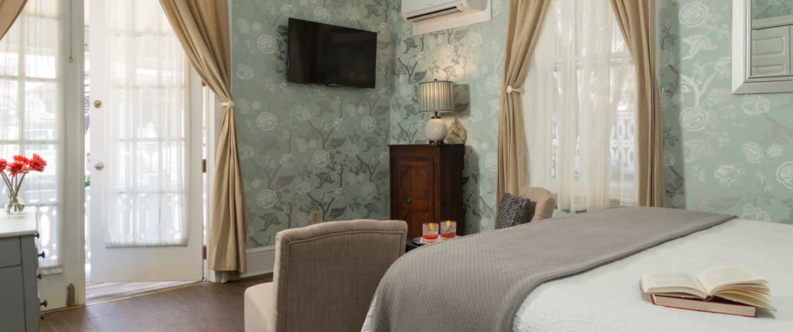 Bedroom with white bedding, gray blanket, sitting area with tan upholstered chairs, and wall-mounted flat-screen TV