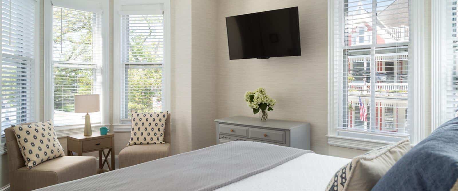 Bedroom with white bedding, gray blanket, sitting area with tan upholstered chairs, and wall-mounted flat-screen TV