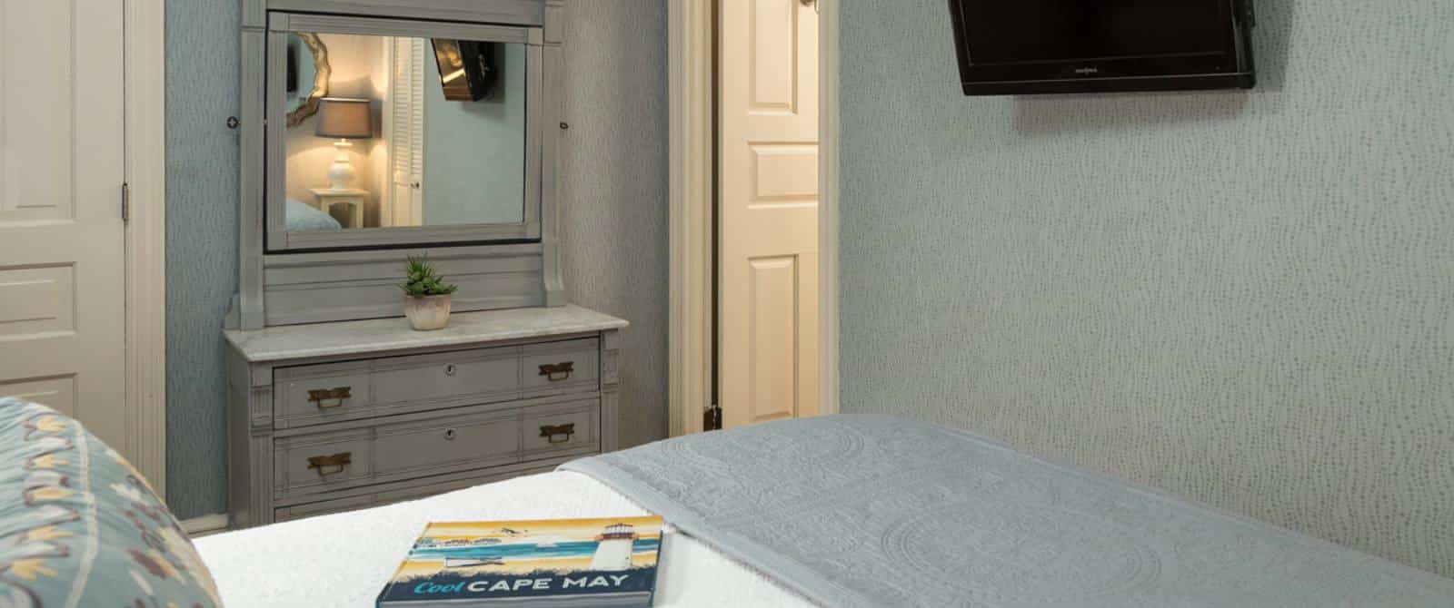 Bedroom with white bedding, gray dresser with mirror, light gray patterned wallpaper, and wall-mounted flat-screen TV