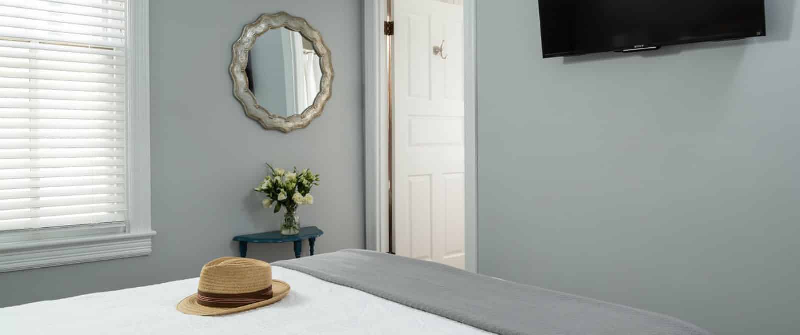 Bedroom with white bedding, gray blanket, light gray walls, mirror on the wall, and wall-mounted flat-screen TV