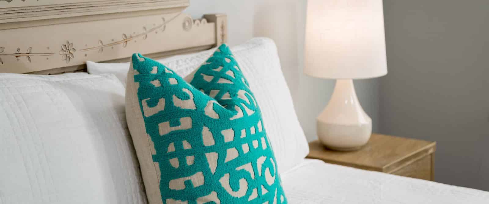 Close up view of bed with ornate headboard, white bedding, teal and tan pillow, and light-wooden nightstand