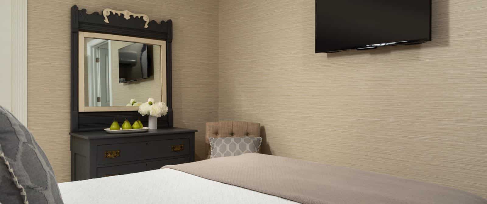 Bedroom with dark wooden dresser and mirror, white bedding, gray blanket, light-tan and modern wallpaper, and wall-mounted flat-screen TV