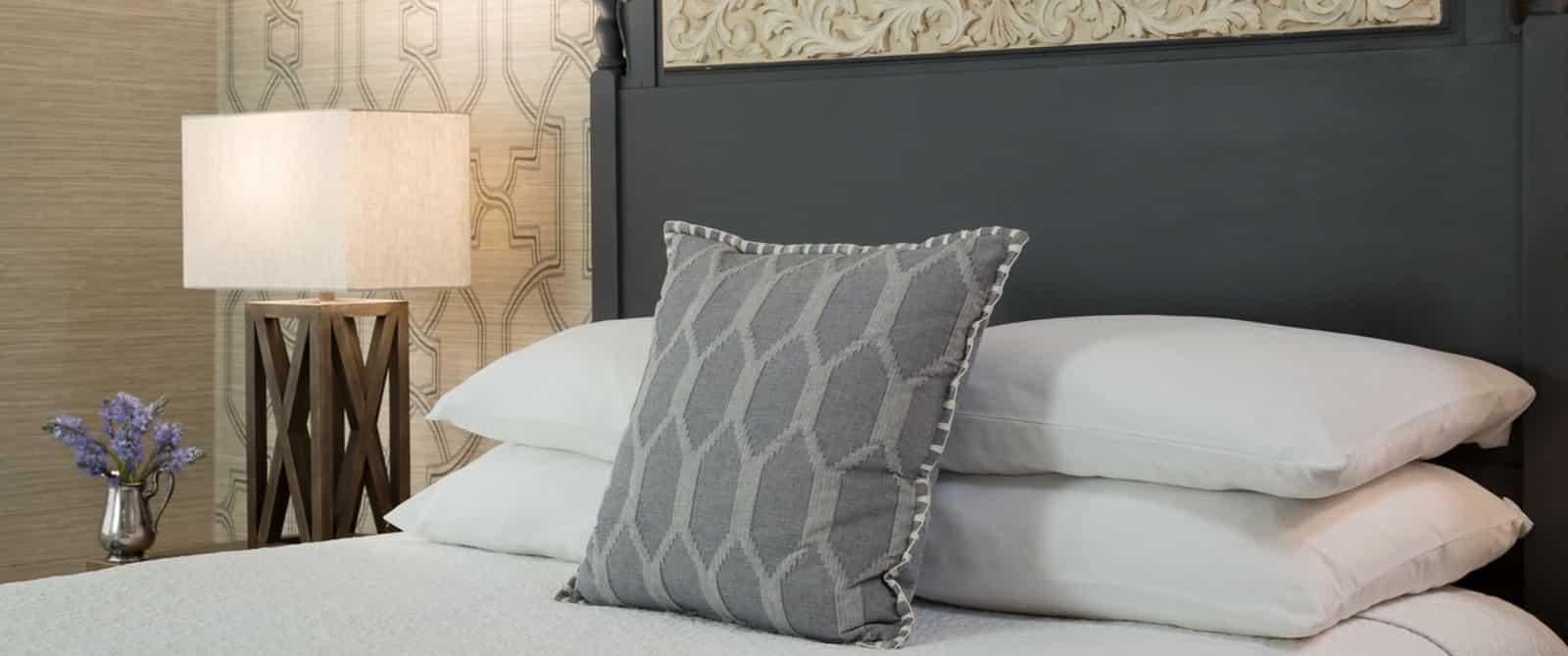 Close up view of bed with dark wooden headboard, white bedding, gray pillow, and wooden lamp