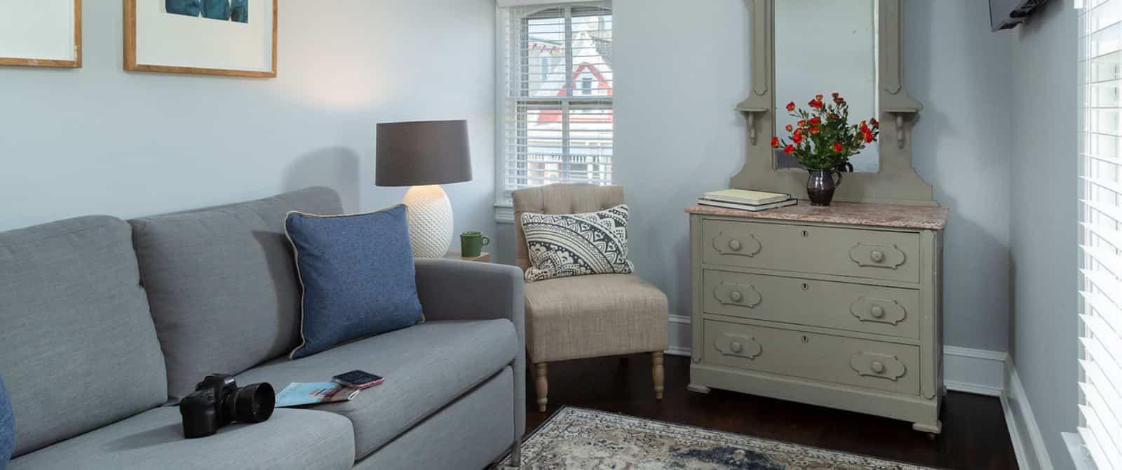 Sitting area with gray upholstered couch, digital SLR camera, denim pillow, hardwood flooring, light gray walls, and gray-painted dresser with mirror on top