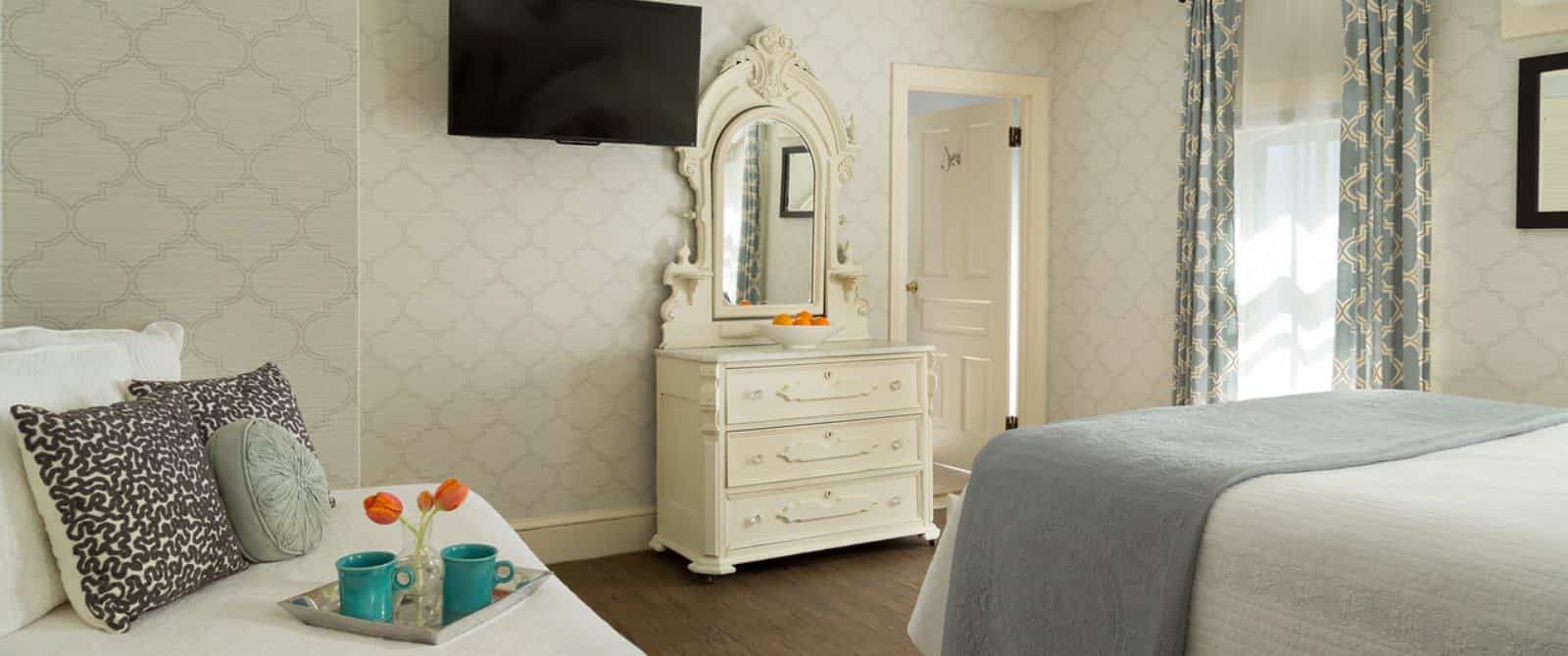 Bedroom with light wooden ornate headboard, matching dresser with mirror, daybed, white bedding, gray blanket, hardwood flooring, and wall-mounted flat-screen TV