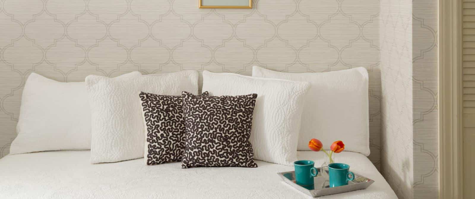 Close up view of a daybed with white bedding, light colored wallpaper, and small silver tray with two turquoise mugs and vase with orange tulips