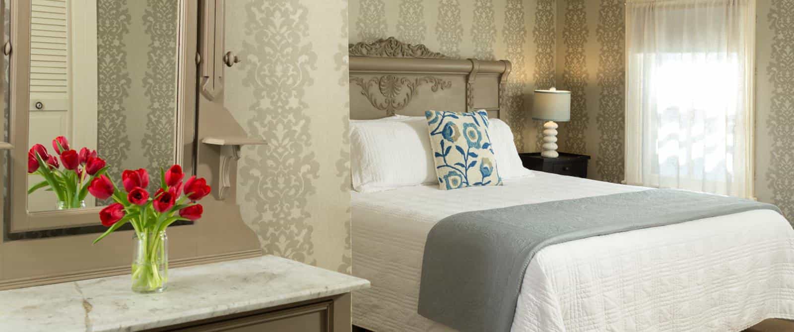 Large bedroom with gray wooden ornate headboard, matching dresser with marble top and mirror, white bedding, hardwood flooring, and brocade wallpaper