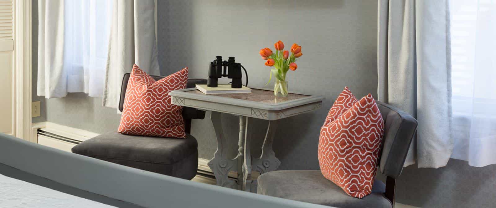 Sitting area with gray upholstered chairs, orange and white brocade pillows, antiqued table, binoculars, and small vase with orange tulips