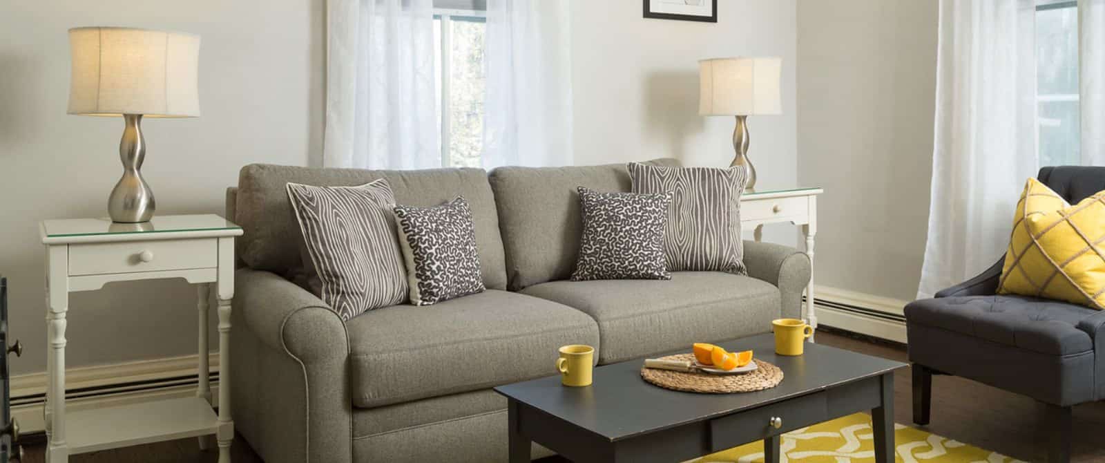 Sitting area with gray upholstered coach, gray and white and yellow pillows, hardwood flooring, light walls, dark wooden coffee table and yellow and white area rug