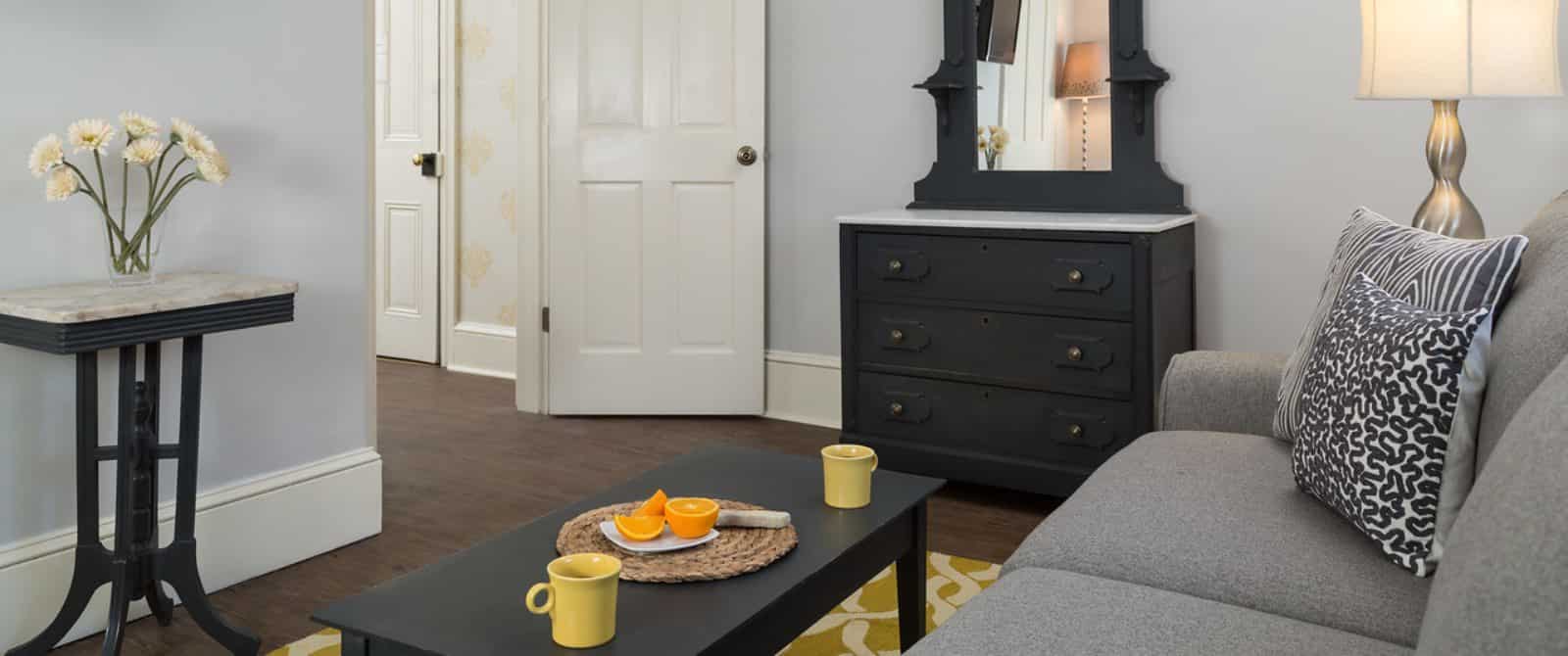 Sitting area with gray upholstered coach, gray and white pillows, hardwood flooring, light walls, dark wooden coffee table and yellow and white area rug