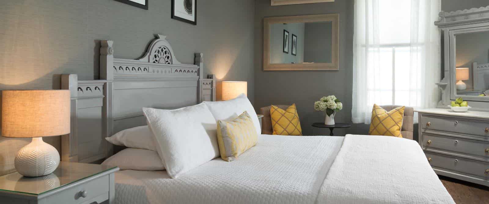 Bedroom with large gray headboard, white bedding, yellow pillows, hardwood flooring, gray walls, and gray dresser with marble top and mirror