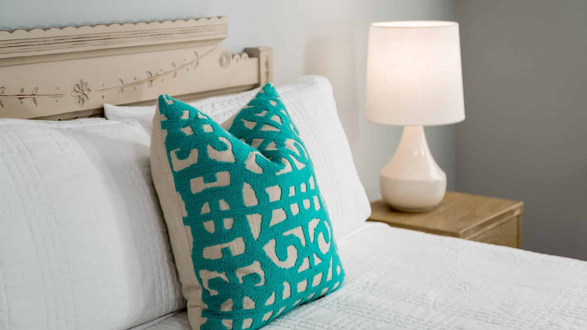 Close up view of bed with ornate headboard, white bedding, teal and tan pillow, and light-wooden nightstand