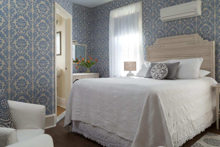 Bedroom with light-colored ornate headboard, white bedding, hardwood flooring, brocade blue wallpaper, and sitting area with white slipcovered chairs