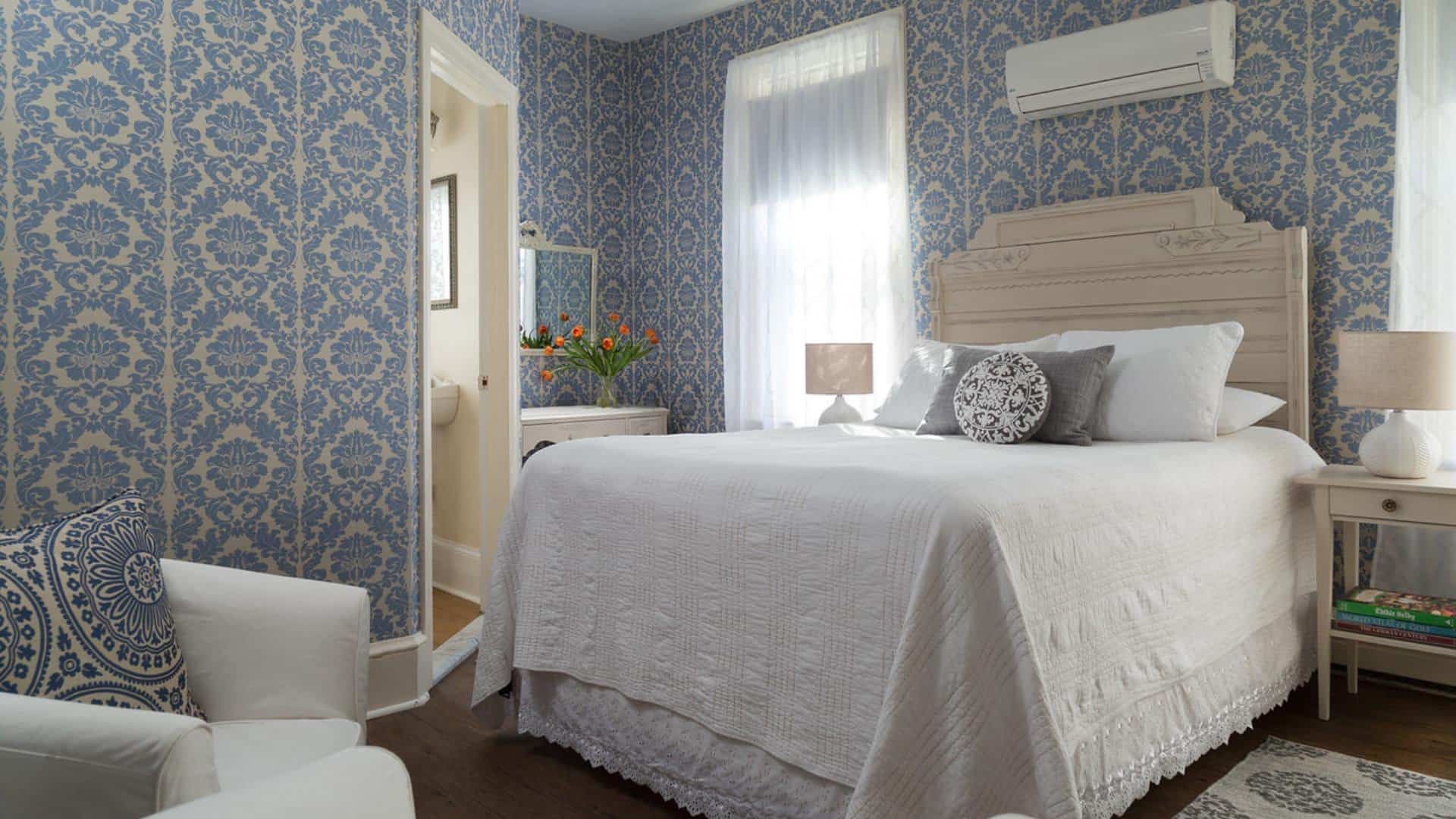 Bedroom with light-colored ornate headboard, white bedding, hardwood flooring, brocade blue wallpaper, and sitting area with white slipcovered chairs