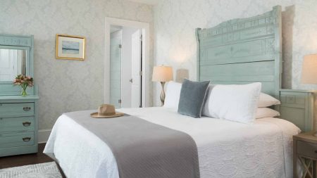 Bedroom with large ornate seafoam headboard, matching dresser with mirror, white bedding, gray blanket, hardwood flooring, and light brocade wallpaper